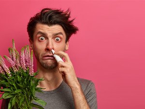 Mann zeigt Allergiesymptome und hält Blumen und ein Nasenspray in der Hand