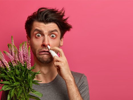 Mann zeigt Allergiesymptome und hält Blumen und ein Nasenspray in der Hand